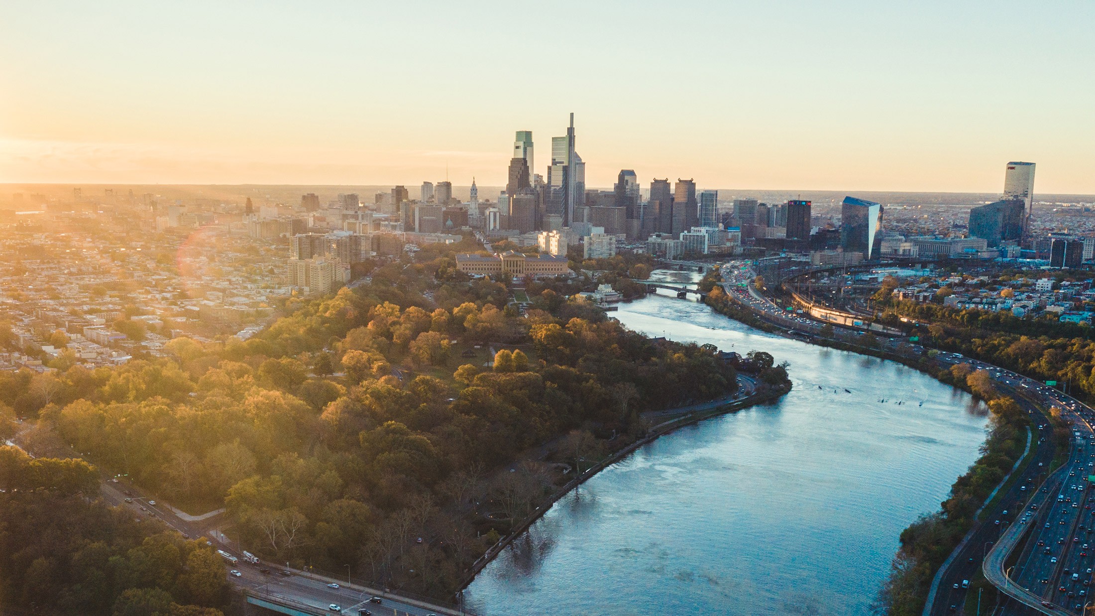 Aerial view of Philadelphia at golden hour, the Schuylkill curving past Fairmount Park toward the Center City skyline
